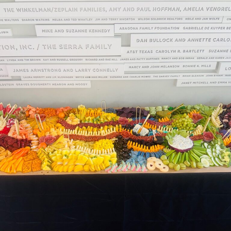 Long rainbow grazing table in front of a donor wall, ZACH Theatre